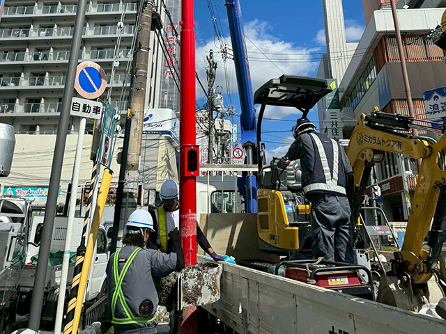 都市部の晴天下で、ヘルメットと空調服を着用した作業員たちが、作業を行っている様子。