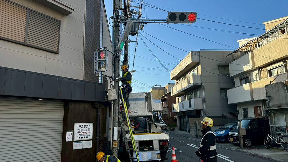 住宅街の交差点で、夕方の光を背景に歩行者用信号機のメンテナンスを行う現場
