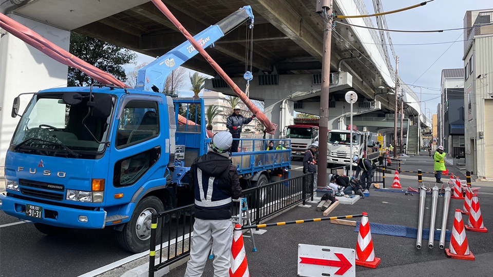 高架下の道路脇で、青いクレーン車を使用してオレンジ色の保護カバーがついた支柱を吊り上げている様子