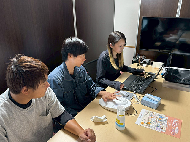 オフィスにて、男女の若手スタッフがリラックスした表情でノートパソコンを囲み、現場情報の共有や打ち合わせを行う様子。