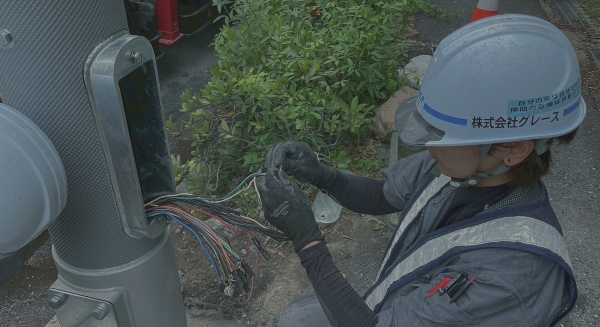 ヘルメットを被った作業員が、信号機支柱のメンテナンス口を開け、中の色とりどりの配線を一本ずつ丁寧に接続・整理している手元のアップ。