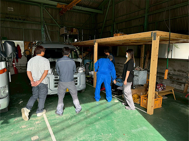 整理されたガレージ兼倉庫にて、作業車（軽バン）のエンジンルームを覗き込み、出発前の点検を行うチームの日常。