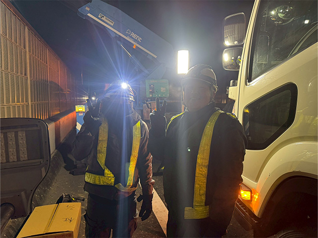 夜間工事の合間に、反射ベストとヘッドライト姿で笑顔のピースサインを送る2人の作業員。職場の仲の良さが伝わるオフショット。