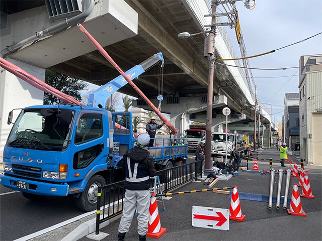 クレーン車で重い支柱を吊り上げ、地上スタッフと合図を送り合いながら慎重に設置を進めるチーム施工の様子。