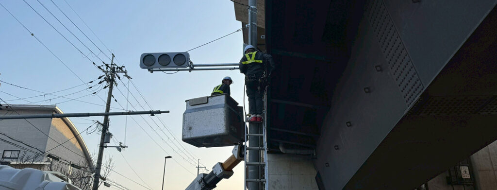 高架下付近の電柱にて、高所作業車と梯子を使い分けて信号機の調整・点検を行う2人の作業員。